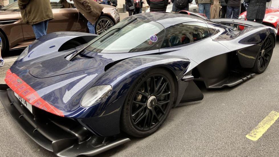 An Aston Martin Valkyrie, a hypercar with curved bodywork and low-profile tyres, in navy and red, parked on Pall Mall