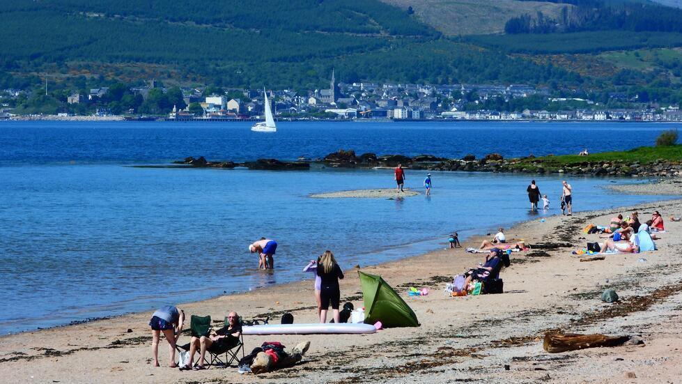 People sit on the sand by the waterside on a sunny day. A town can be seen in the distance across the water