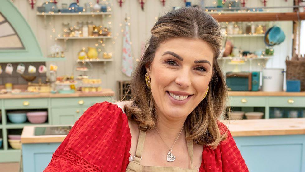 Georgie Grasso smiling at the camera. She is wearing a red top underneath a beige apron. She is stood in the Bake Off tent. It is a head and shoulders shot of her. 