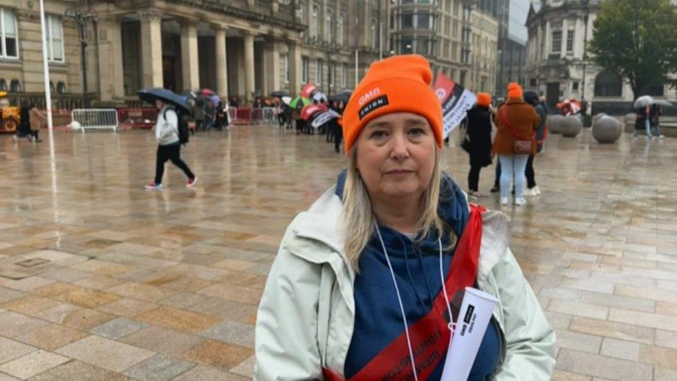 A woman with blonde hair and a cream jacket wearing an orange GMB union hat. In the background people are standing under umbrellas or walking holding umbrellas. 