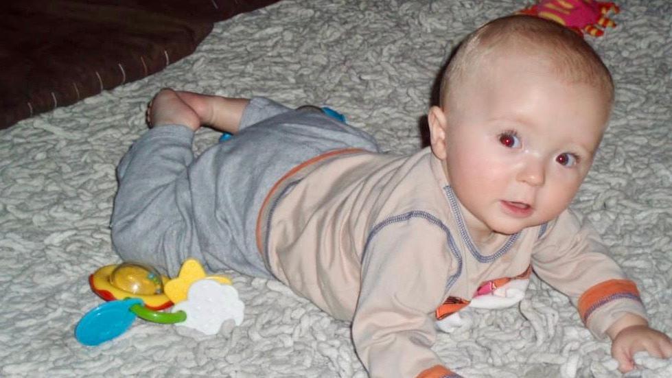 Baby Alex on a white mat with toys around him. He is on his stomach and looking at the camera.