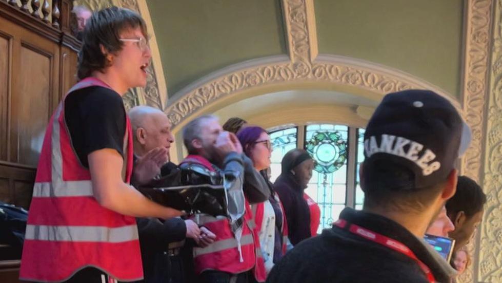 A group of people in high-vis pink and grey jackets, some of them visibly angry and shouting into a space to the right of them.