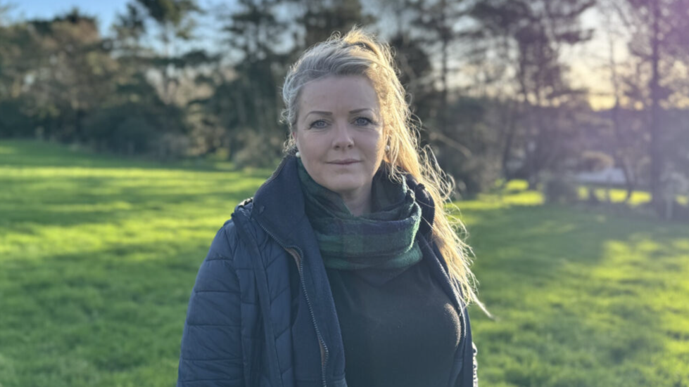 Kirree Kermode is standing in a green field lined with trees on a sunny morning. She has long blonde hair tied up and is wearing a navy coat with a green and blue tartan scarf.