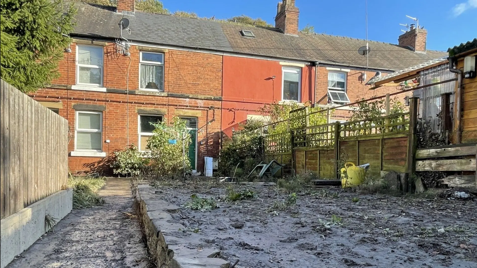A terraced red brick house with a garden covered in mud