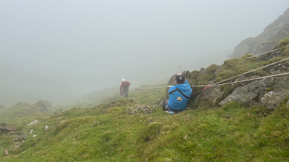 Two men in outdoor gear, on a green mountain side with helmets and ropes.