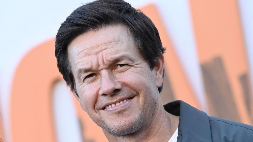 Mark Wahlberg has dark hair and is wearing a grey jacket over a white T-shirt. He is smiling.