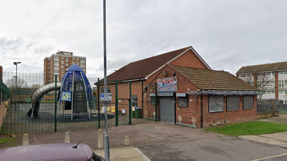 A children's play area and building viewed from the road. There is a large, brick building in the middle-right of the photo, with one smaller building on the front attached to a larger, taller building at the rear. The front building has a grey shutter door and windows, chalk drawings of flowers along the side, barbed wire along the edge of the roof, and graffiti writing above the door. To the left there is a metal blue and grey children's climbing frame. A tower block is visible in the background, set against a grey, lightly cloudy sky.
