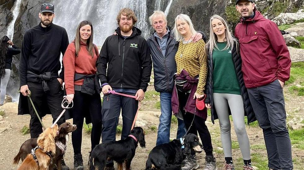 A row of seven people in front of a waterfall.  Four are holding dogs on leads. Alex with short hair and beard and Trevor with short white hair are in the group.