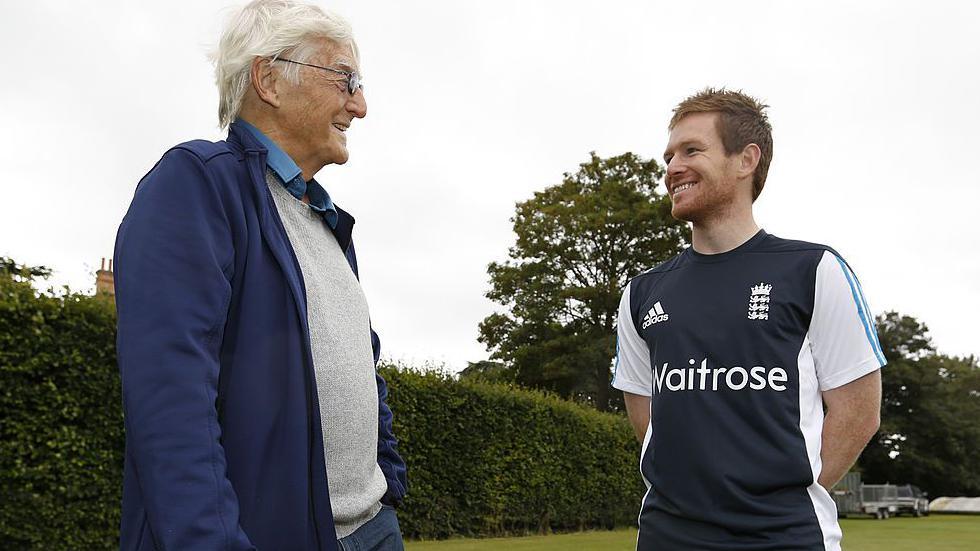 Sir Michael Parkinson, wearing a blue jacket, grey jumper and light blue shirt under it, talking to Eoin Morgan, dressed in an England cricket top, in front of a tree and a hedge at the cricket club 