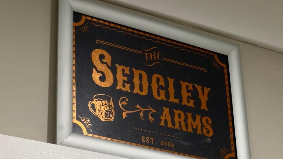A black and gold plaque which says the Sedgley Arms, est 2020. There is also a beer glass in gold. 