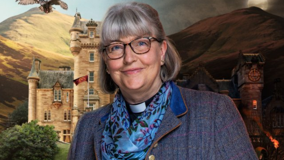 The Reverend Lisa Coupland is wearing a scarf, jacket and glasses in front of a backdrop of a castle and mountains.