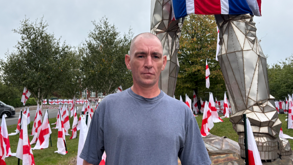 A man in a grey T-shirt and a bald head stands next to a large silver statue that has a union jack flag draped over it. There are dozens of St George's flags pressed into the ground around him. 