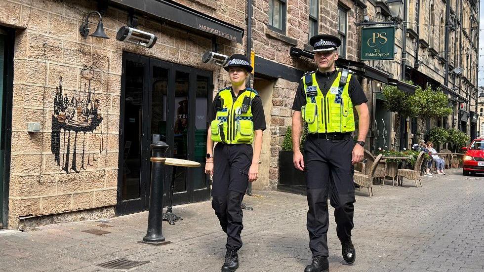 Two police officers, one male and one female wearing high-visibility jackets, walking past The Pickled Sprout restaurant in Harrogate.