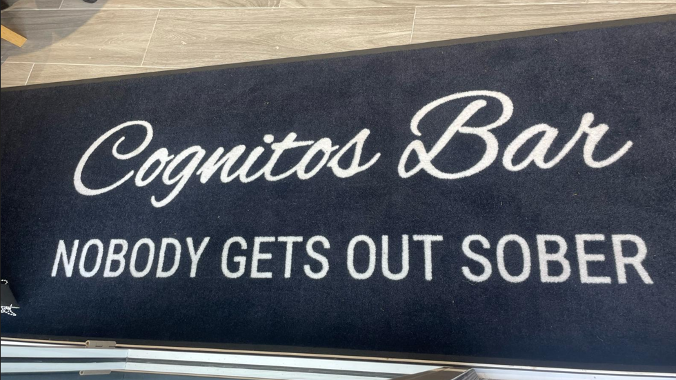 A navy mat shows the words "Cognitos Bar - Nobody gets out sober" and is on the floor of the entrance to the bar.