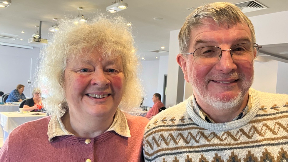 Helen is wearing a peach pink cardigan with golden buttons and Graham wearing a crème coloured winter jumper. 