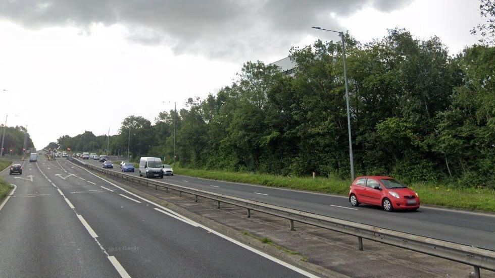 Cars drive along a motorway. There are two lanes on each side of the central reservation, trees can be seen on the far right. 