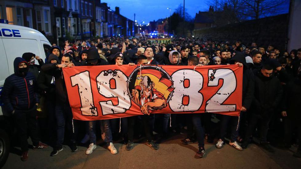 A large crowd of Atletico Madrid fans are kettled as they walk together down a street to Anfield. The fans at the front hold a large red banner with the number 1982 emblazoned on it.