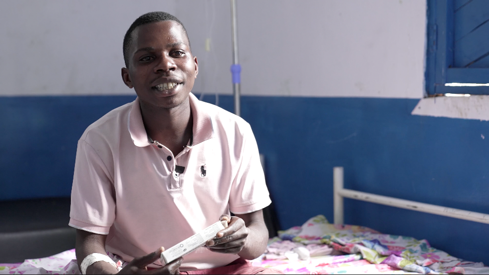 Sadiki Bichichi Aristide sits on a hospital bed, holding a medication packet