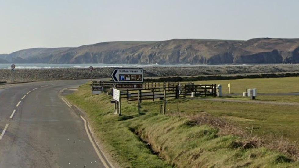 A coastal road, with a field on one side and a shingle bank on the other