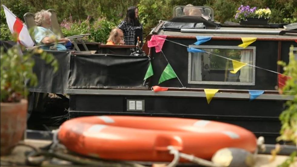 Three people on a canal boat decorated with bunting, also flying an England flag