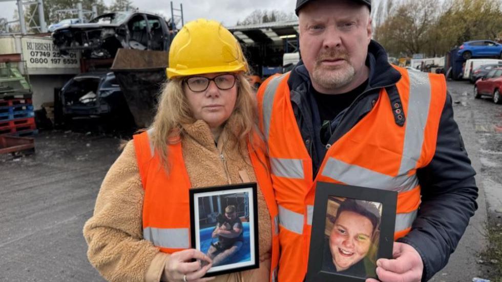 A woman with long blond hair, a beige coat, an orange reflective jacket and a white safety helmet holding a photo of a boy next to a man with grey hair and a beard in a black cap and coat and an orange reflective jacket on an open space with a number of wrecked cars standing in piles
