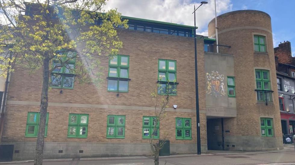 Luton Crown Court: A tree with yellow leaves stands to the left of a brown-brick building with green-framed windows. The words "CROWN COURT" are written above the entrance of the building.