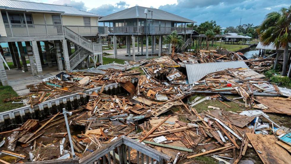 Photograph of a couple of houses on stilts and a lot of debris close by resulting in damage from a hurricane.