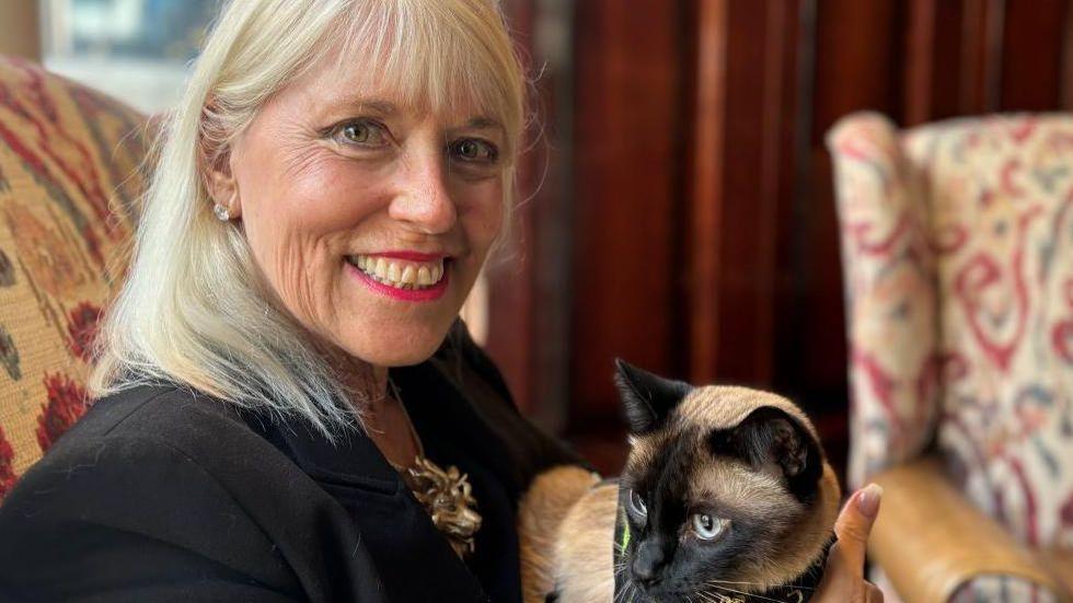 Holly Hennessy, a woman with shoulder-length grey-blonde hair, wearing a black jacket, holds her pet cat captain. The cat is tan and black in colour.