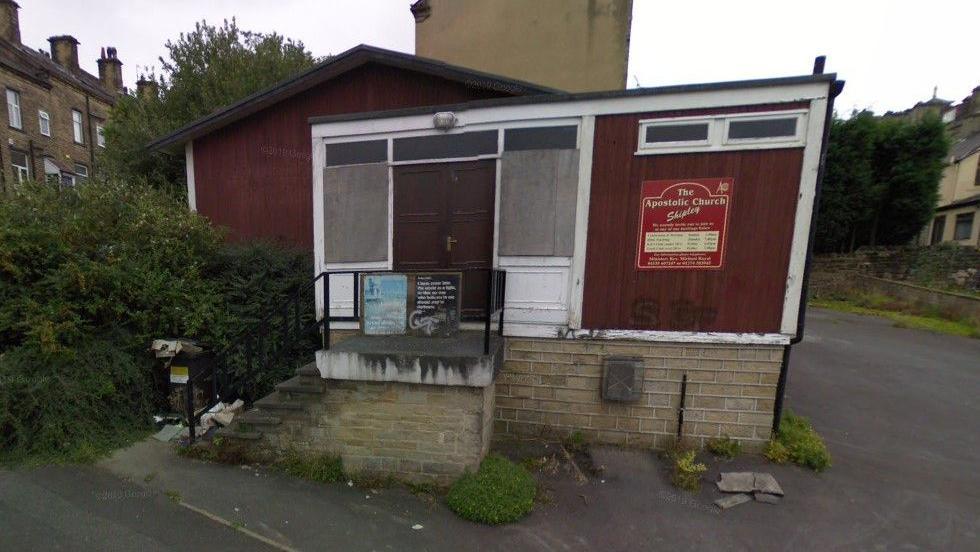 An old church building, featuring a sign on the outside wall indicating its use and a small set of steps leading up to a pair of brown double doors.