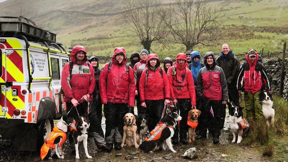 Mountains rescuers in red jackets and black trousers standing at the bottom of a peak in the rain. They have rescue search dogs with them. A van is parked on the left and there is a stone fence to the right. 