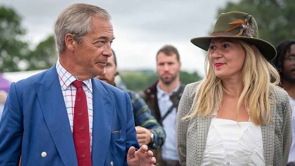 Leader of Reform UK Nigel Farage is on the left, he looks to his right and has a blue jacket on over a white striped shirt and a red tie. He has short grey hair. Laura Anne Jones is stood to the right of him and has a white shirt with a checkered green jacket on. She has shoulder-length blonde hair and a green velvet hat on with a feather. 