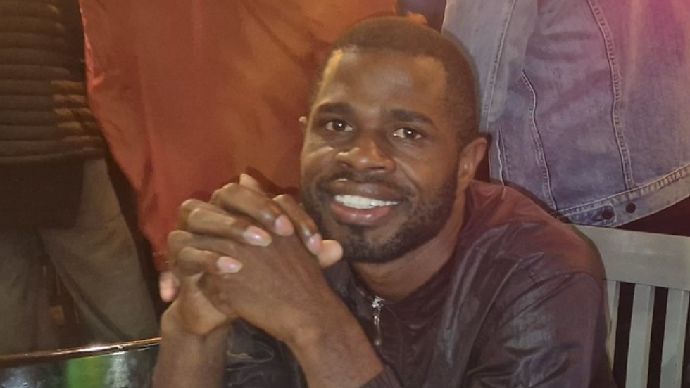 Yostin Mosquera interlocking his hands on a table, smiling and looking at the camera. He is sat down on a chair, and there are several people standing up behind him.