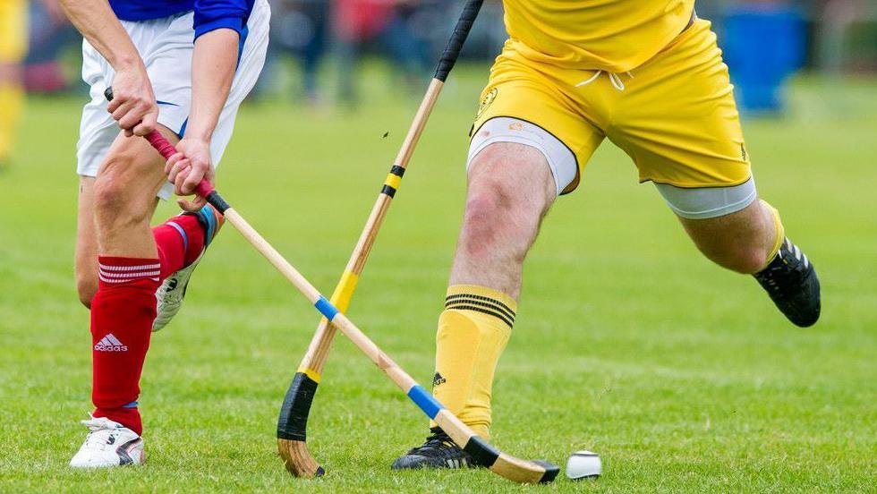 The legs of two shinty players - one of them in blue and white kit and the other in yellow - compete for a ball.