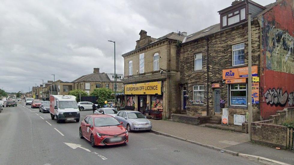 Street View image showing traffic passing by shops including one with a large sign reading European Off Licence