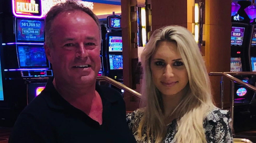 A head and shoulders image of a smiling couple standing in front of rows of upright gambling machines. He is wearing a black v-necked top and has thinning brown hair. She has blonde long hair and is wearing a snakeskin-print blouse
