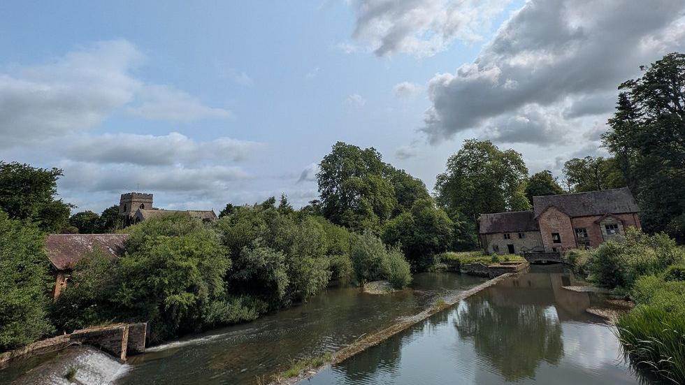 A river leads up to a brick house with a series of large green trees around it which are reflected in the river.