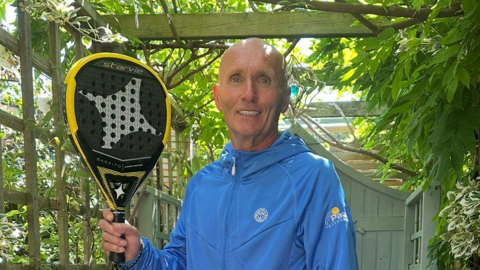Mr Jordan is holding a padel racquet in his right hand. It has been lifted upwards so the camera can see its solid centre. He is standing in what appears to be a garden while wearing a blue jacket with Padel Peterborough branding on. 