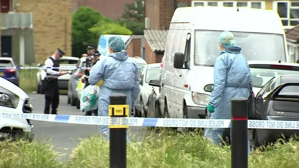 Police tape in the foreground and two people dressed in light blue forensic suits and in the background there are police officers in uniform. There are cars parked on the right handside and buildings in the background.