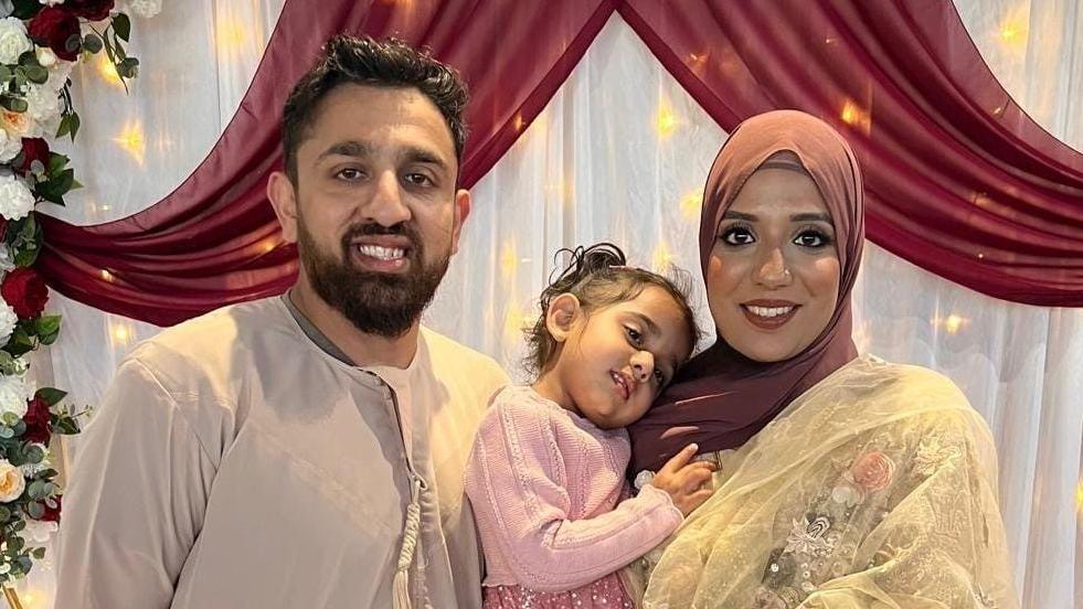 Akeel Nanabawa and Hannaa Vorajee hold their daughter Sara at a celebration event. Akeel wears a grey top while Hanaa wears a terracota head scarf and gold top. Sara is dressed in pink and they stand in front of a floral display