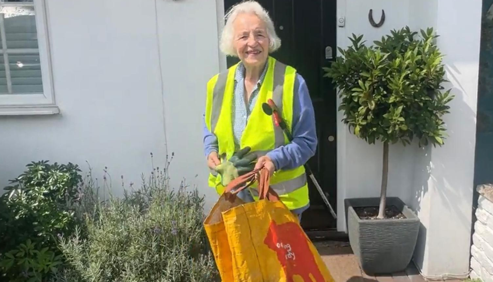 An 85-year-old woman wearing a yellow tabard, holding a litter picking stick and large orange canvas bags.