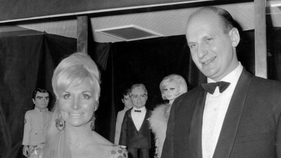 A black and white photo of a bald man wearing a suit and bow tie and a blonde woman wearing a dress and large earrings.