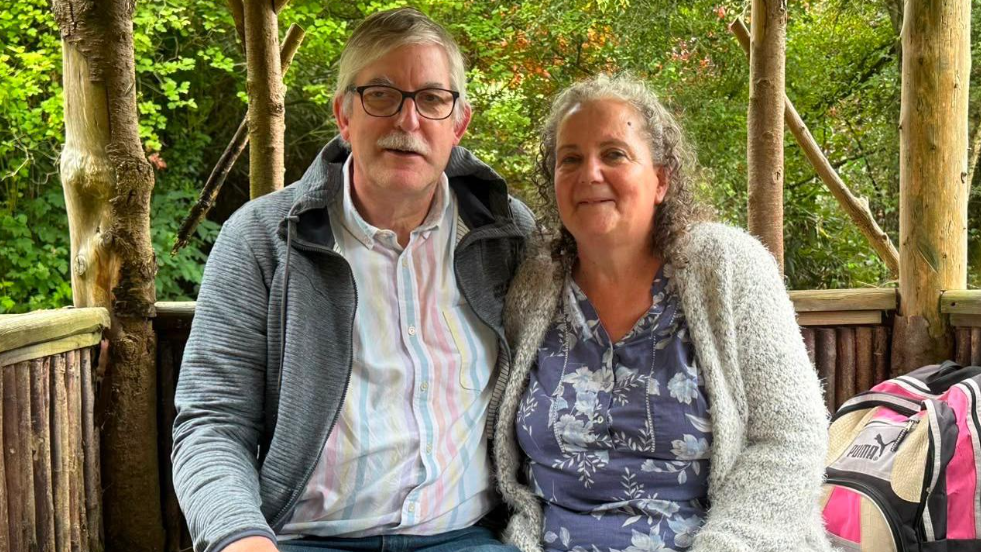 Alan a Julia Dutton yn eistedd mewn man cysgodol o bren gyda choed yn y cefndir