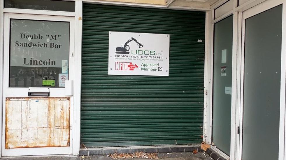 A closed, green shutter at the former shopping centre carries a sign for a demolition firm, with a bulldozer logo. To the left is the door to the former "Double M Sandwich Bar". A panel on the door looks worn and rusty.