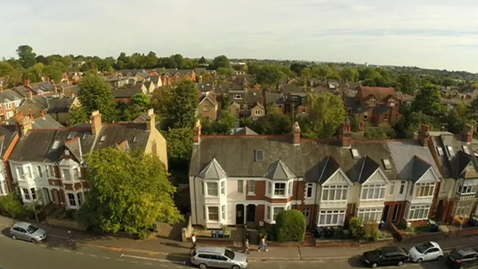 Footage taken from a drone shows a street in Oxford. It has Victorian terraced houses on it. There are several cars parked in front and three people walk along the street, from left to right of the picture. More houses can be seen in the background, along with trees.