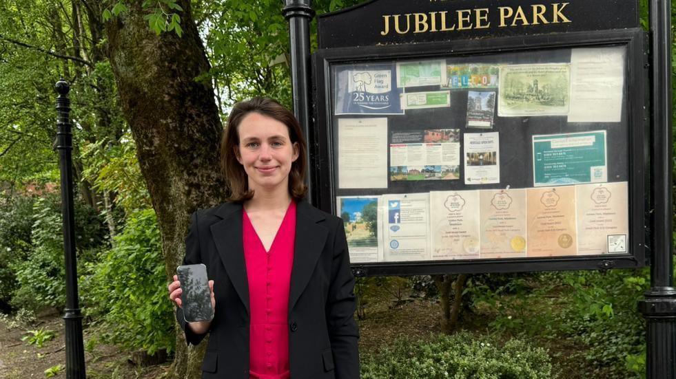 Elsie Blundell, wearing a black blazer over a red top, holds up her mobile phone. She is seen standing in front of a large noticeboad and sign for Jubilee Park