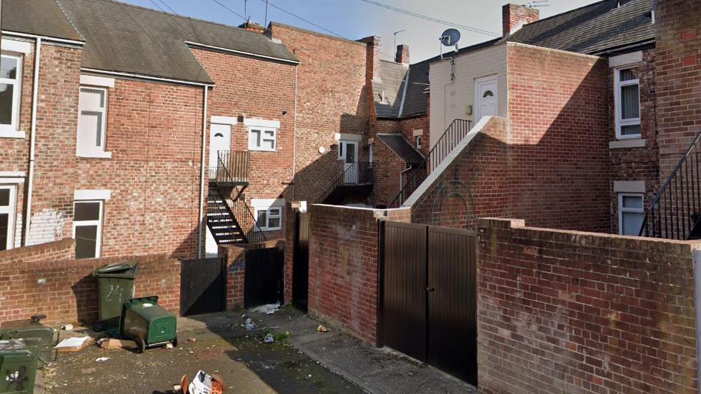 Street view of the back of a row of red brick terraced houses with backyards surrounded by brick walls and black gates. Several of the two-storey houses are actually divided into flats with metal staircases descending from first-floor white doors. Several green wheelie bins are lying on their side