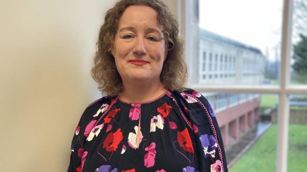 Louise Wallace, director of public health North Yorkshire, pictured wearing a floral dress and standing in front of a window through which can be seen grass and a modern office building