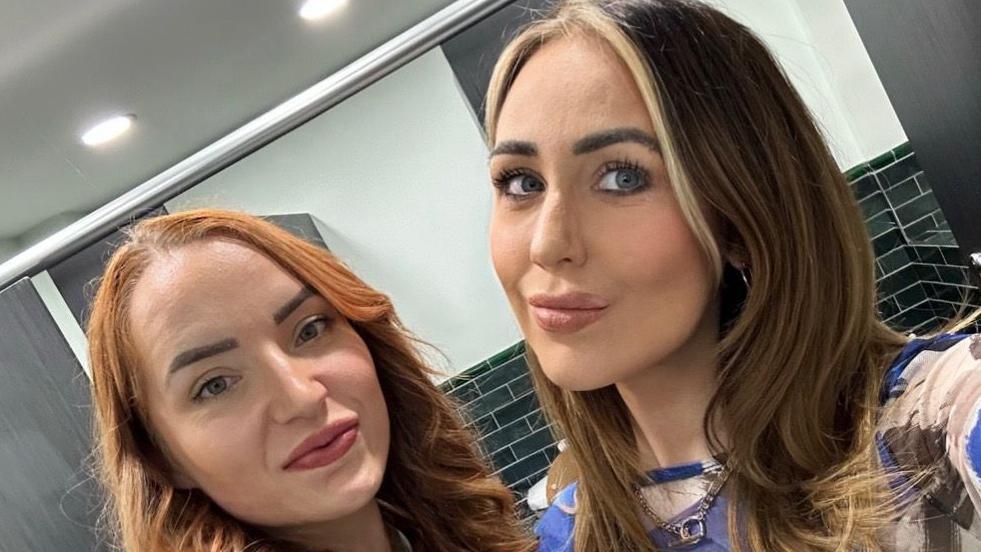 Ashleigh and April are stood taking a selfie next to each other in a bathroom. Ashleigh has shoulder length ginger hair and is smiling. Ashleigh has shoulder length brown hair with blonde bangs, she is smiling at the camera and wearing a blue and cream coloured top.