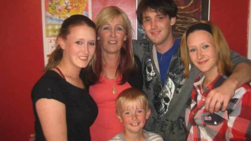 A photo of a family embracing each other. Two women in their 20s stand either side of a young man and a slightly older woman, while a small boy smiles in front of them.