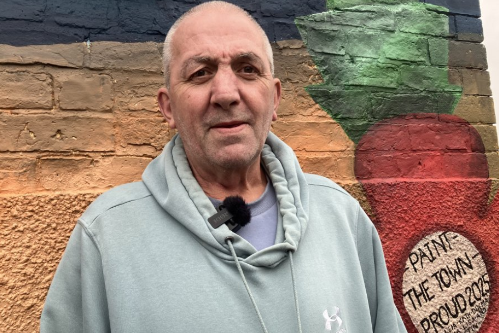 Man with short grey hair wearing a pale blue hoodie standing in front of a mural which is painted on the side of a wall. The wall is orange and grey and has part of a painted red poppy with the words: "Paint the town proud 2025". 